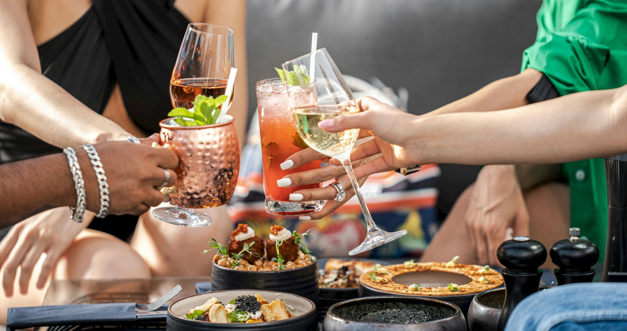 A group of friends cheers with assorted cocktails over a table of tapas and small plates at a lively outdoor get-together.