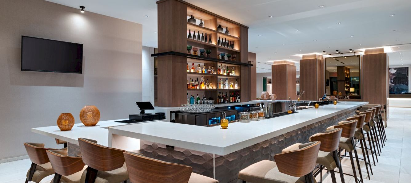 A modern hotel bar with a long white-topped island, wooden stools, shelving stocked with bottles, and a wall-mounted TV in a bright, open space. The bar area is lit warmly.