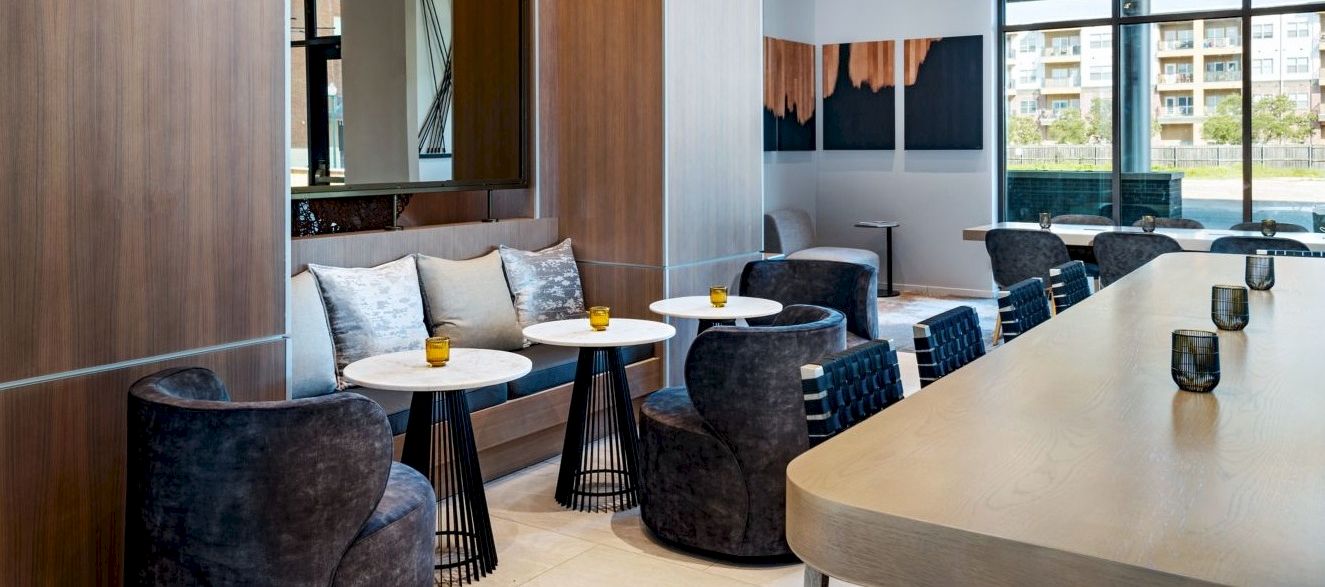 A modern lounge area with wood-paneled walls, comfy dark chairs, small round tables with candles, and a long light countertop&mdash;bright, airy, and stylish.