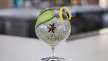 A gin and tonic glass with ice, lime wedge, a star anise garnish, and a yellow straw, set on a bright outdoor table.