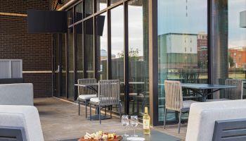 A stylish outdoor dining setup with two gray couches, a small table, wine bottle, and glasses on a plate of snacks, under a glass-walled patio.