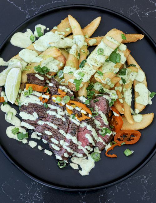 A plate of steak with fries, greens, and a drizzle of sauces atop a dark plate.