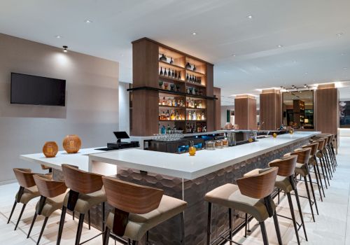 A modern hotel bar with a long white-topped island, wooden chairs, backlit shelves of liquor, a wall-mounted TV, and a bright, airy lounge area.