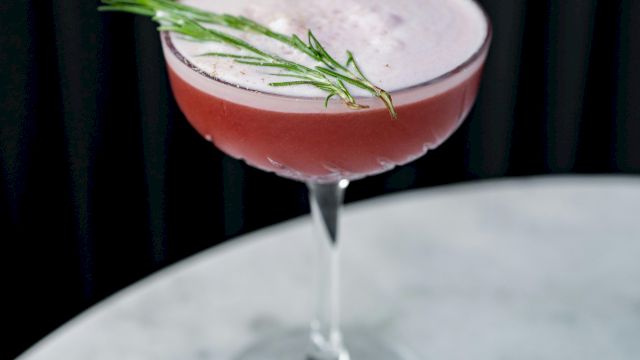 A pink cocktail in a coupe glass with a rosemary sprig garnish on top, set against a dark background.