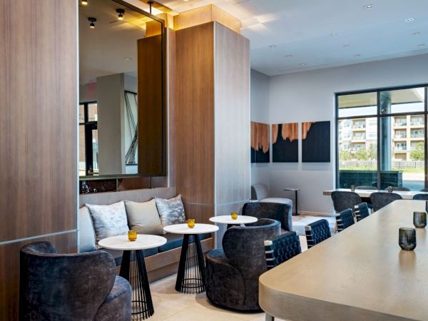 A modern lounge or hotel lobby with a long curved reception or bar counter, wicker-style chairs, small round tables, wood paneling, and large windows.