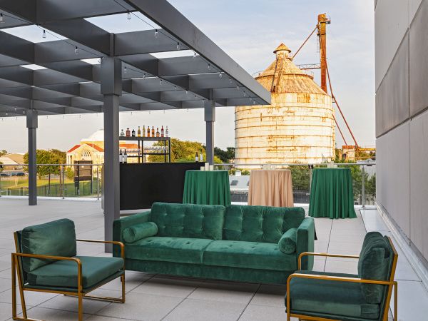 A stylish outdoor seating area with green velvet sofas and chairs, on a rooftop with a historic temple in the background, under a shaded canopy.