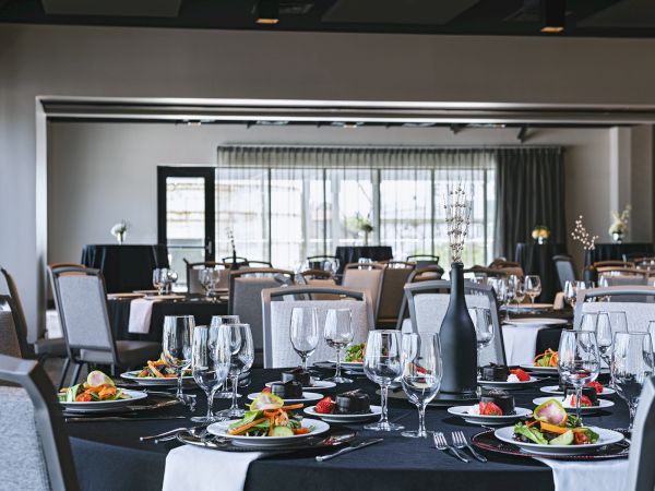 A formal dining room set for a banquet: round tables with black tablecloths, elegant place settings, glassware, plates, and a bright, airy backdrop.