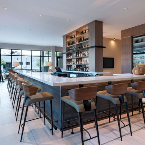 A modern, bright kitchen and dining area with a long wooden table, bar stools, pendant lights, and open shelving along the wall.