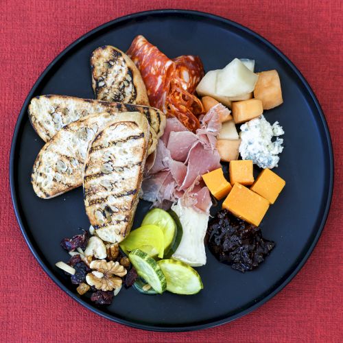 A round charcuterie plate with grilled bread slices, prosciutto, cheddar cubes, blue cheese, olives, walnuts, grapes, melon, cucumber, and crackers on a dark plate.
