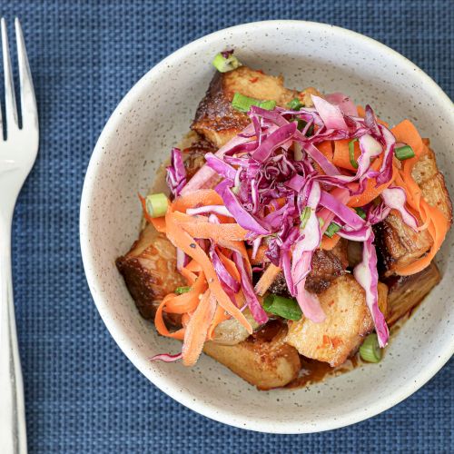 A bowl of colorful tacos or noodles with pink cabbage, shredded veggies, and sauce, plated beside a fork on a dark blue napkin.