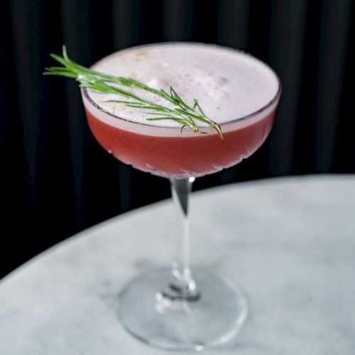 A pink cocktail in a coupe glass with a lime garnish and a frothy top, on a white surface with a dark background.