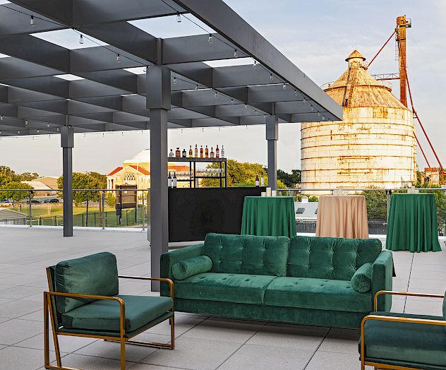 A stylish rooftop seating area with green velvet sofas and chairs, set against a temple-like structure in the background, under a shaded canopy.